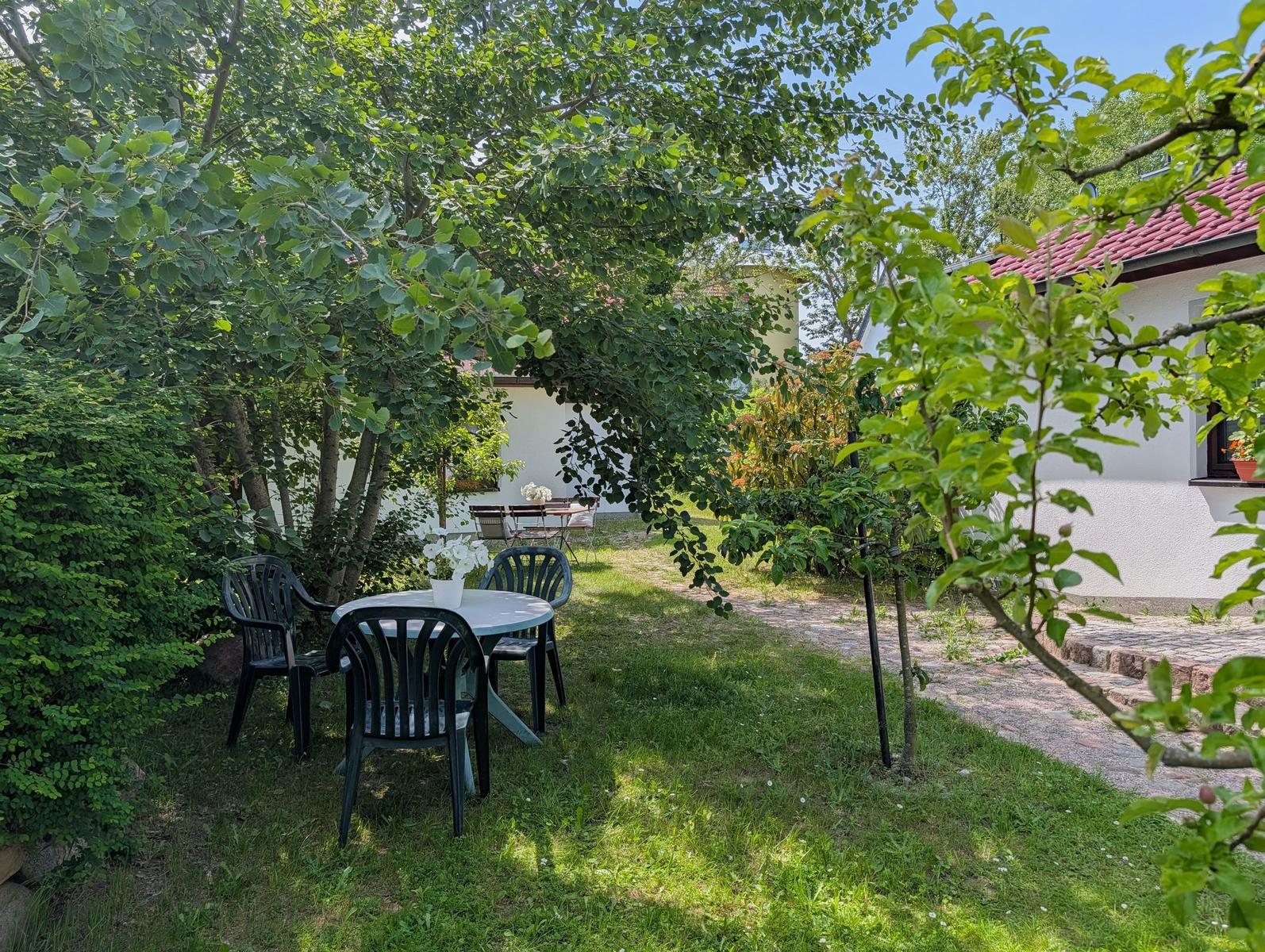 Garten mit Tisch und Stühlen unter Bäumen. Weiße Wand und roter Dachfirst sichtbar.