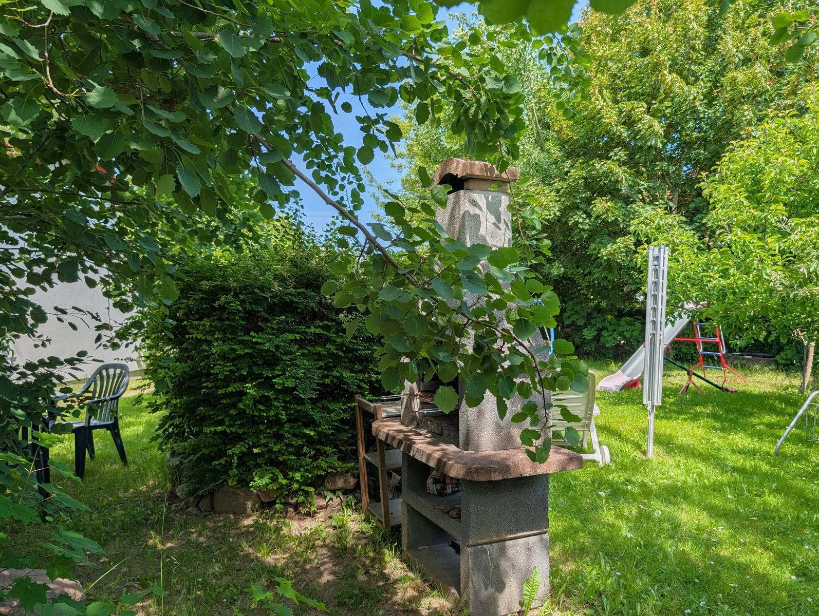Garten mit Grill, Spielplatz und Sitzgelegenheiten unter Bäumen.