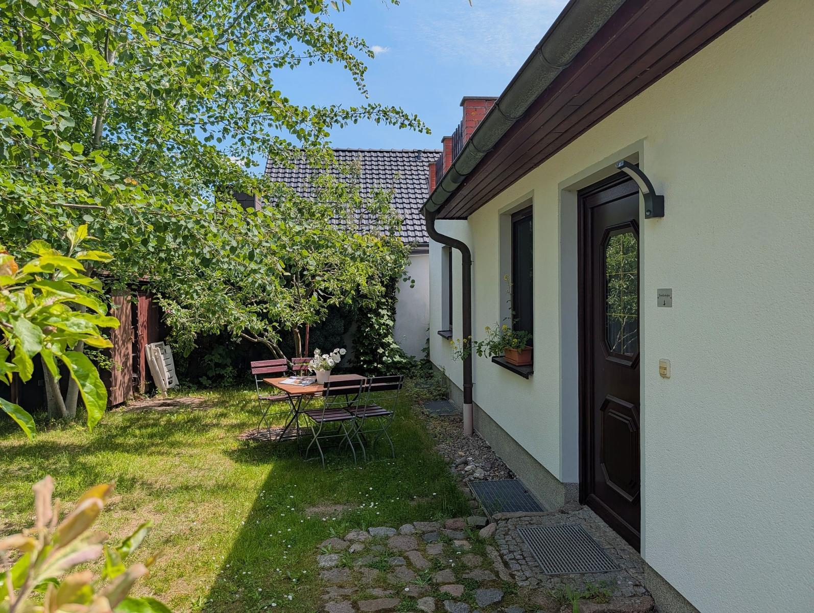 Hinterhof mit Tisch, Stühlen und grünem Rasen. Eingangstür und Fenster mit Pflanzen.