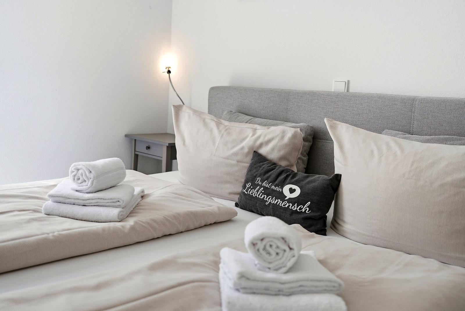The bed is decorated with pillows and towels. A wall lamp and nightstand are visible.