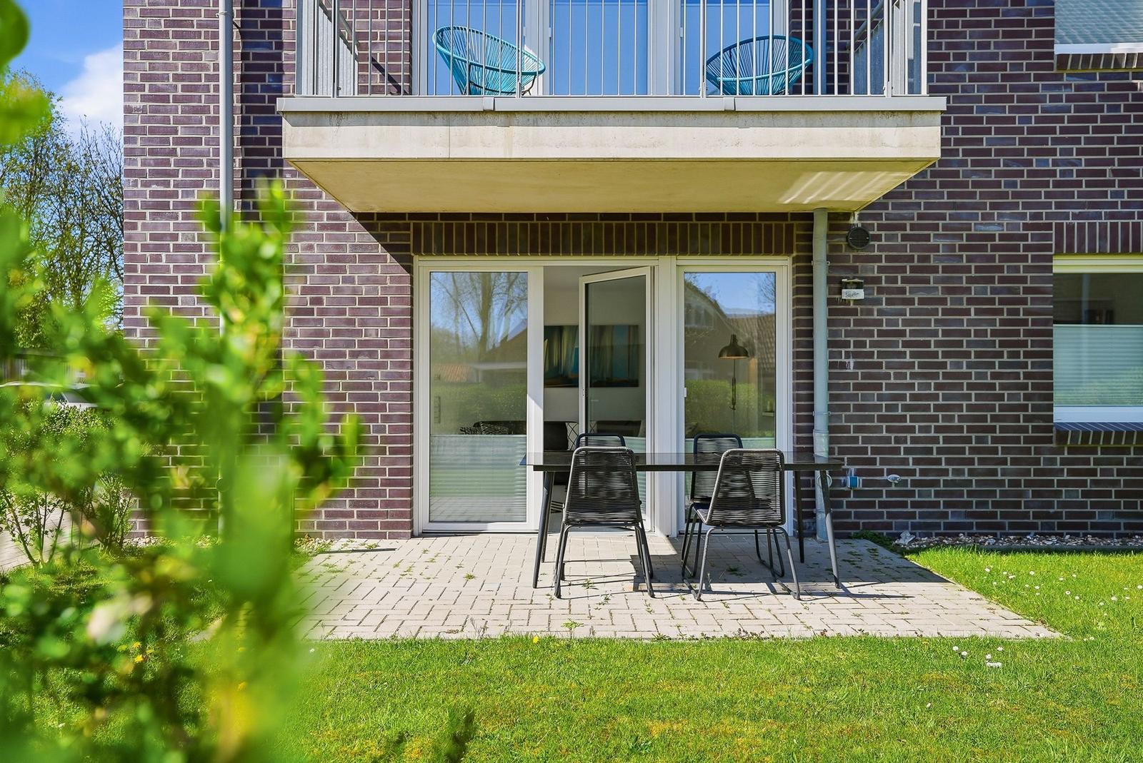 Terrace with table and chairs in front of a house with balcony and glass doors.