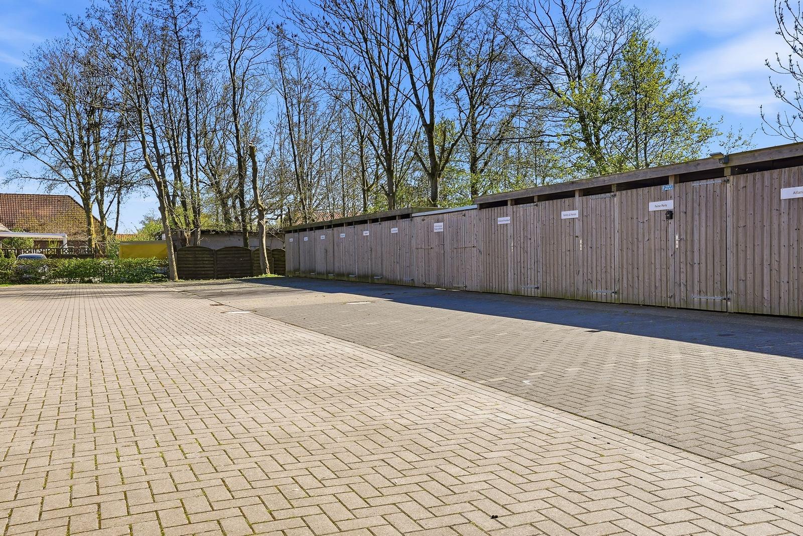 Paved area with wooden garages and trees in the background.