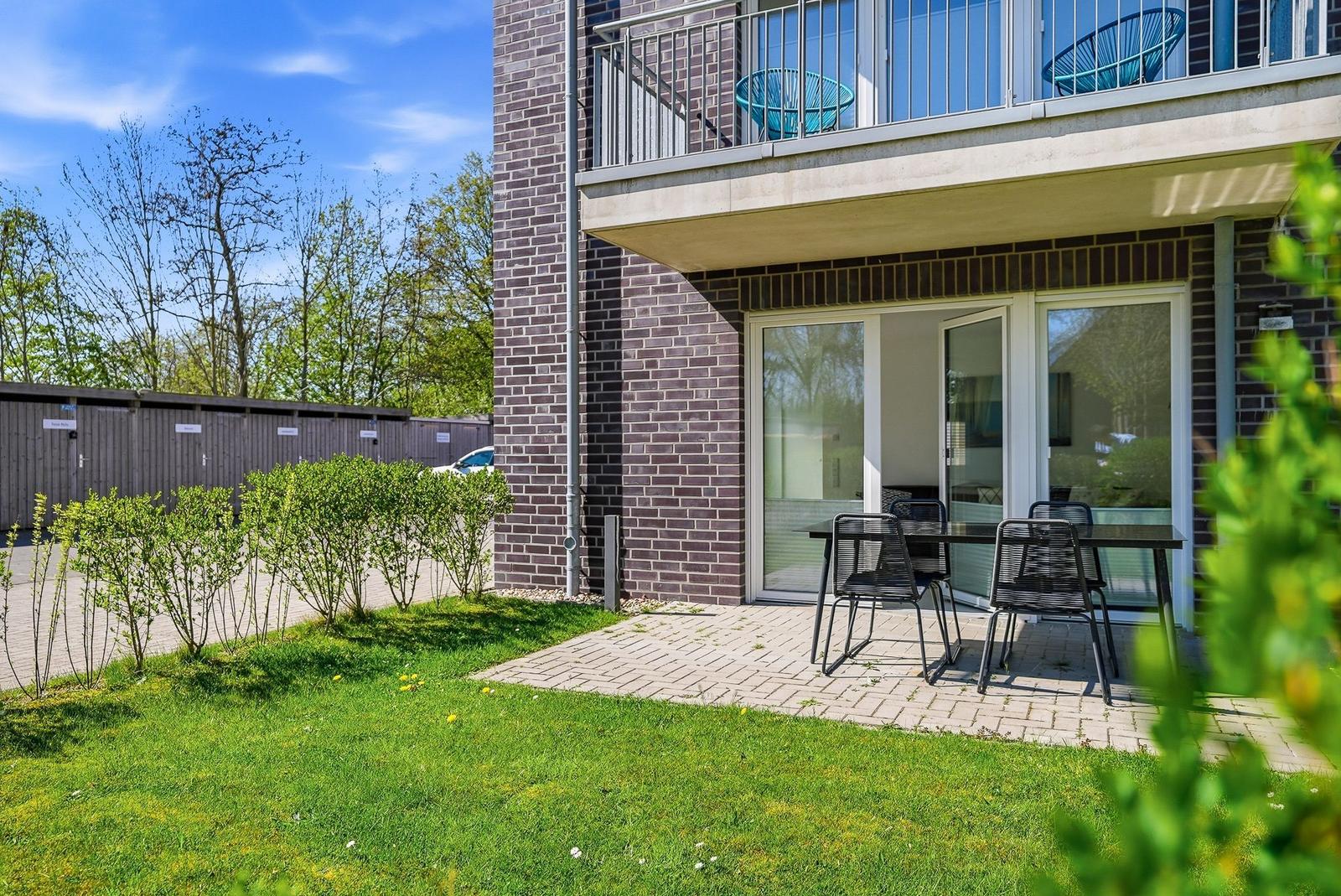 Terrace with table and chairs in front of a building with balcony and glass doors.