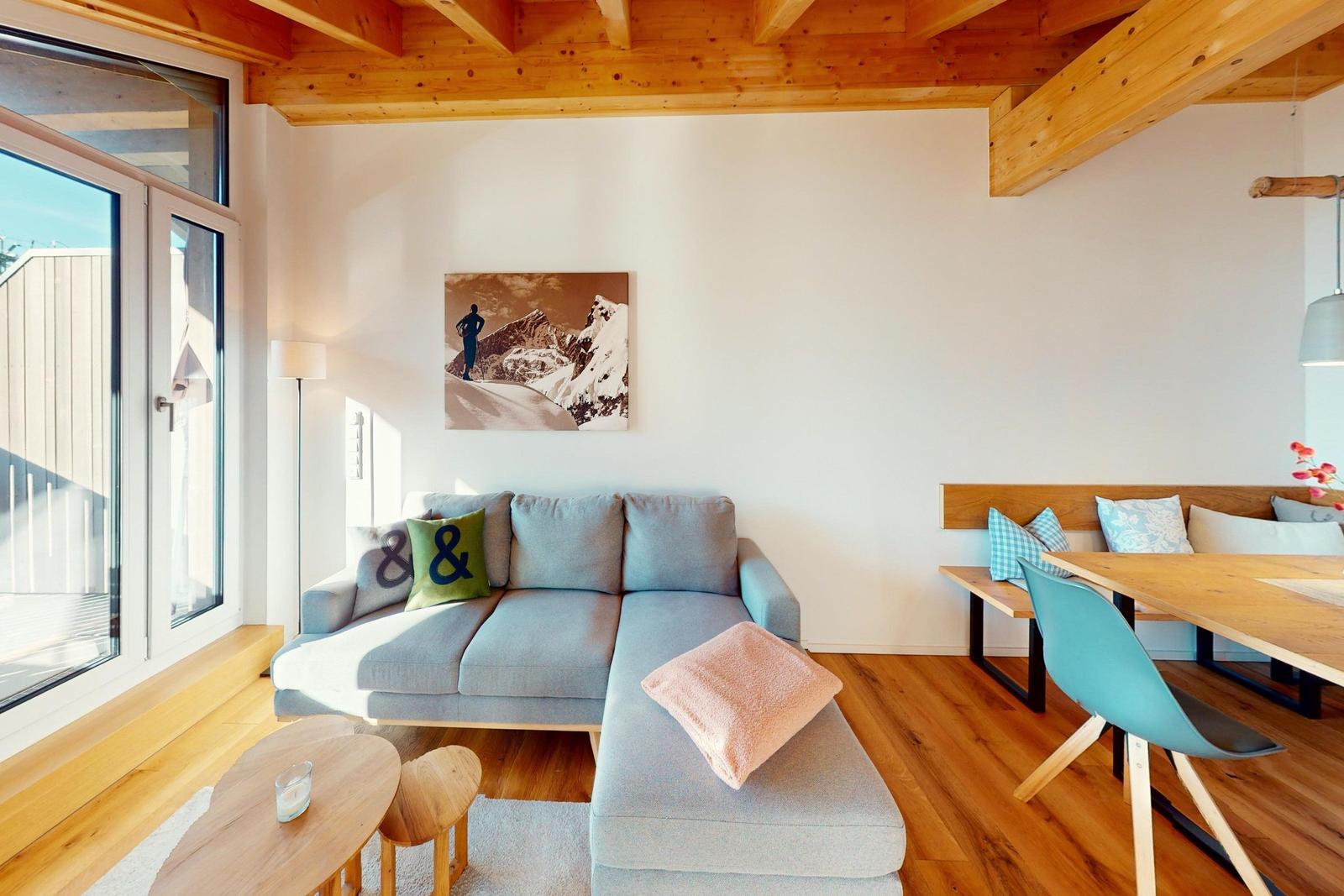 Wohn- und Essbereich mit Sofa, Tisch und Stühlen. Holzdecke und Fenster sichtbar.