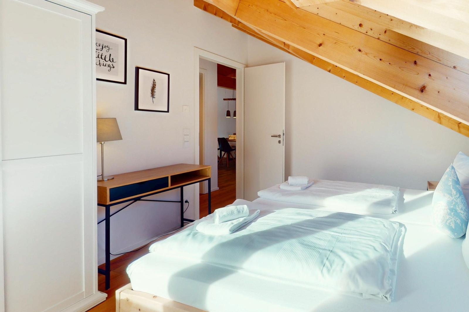 Schlafzimmer mit Doppelbett, Schreibtisch und Holzbalkendecke. Türen führen in angrenzende Räume.