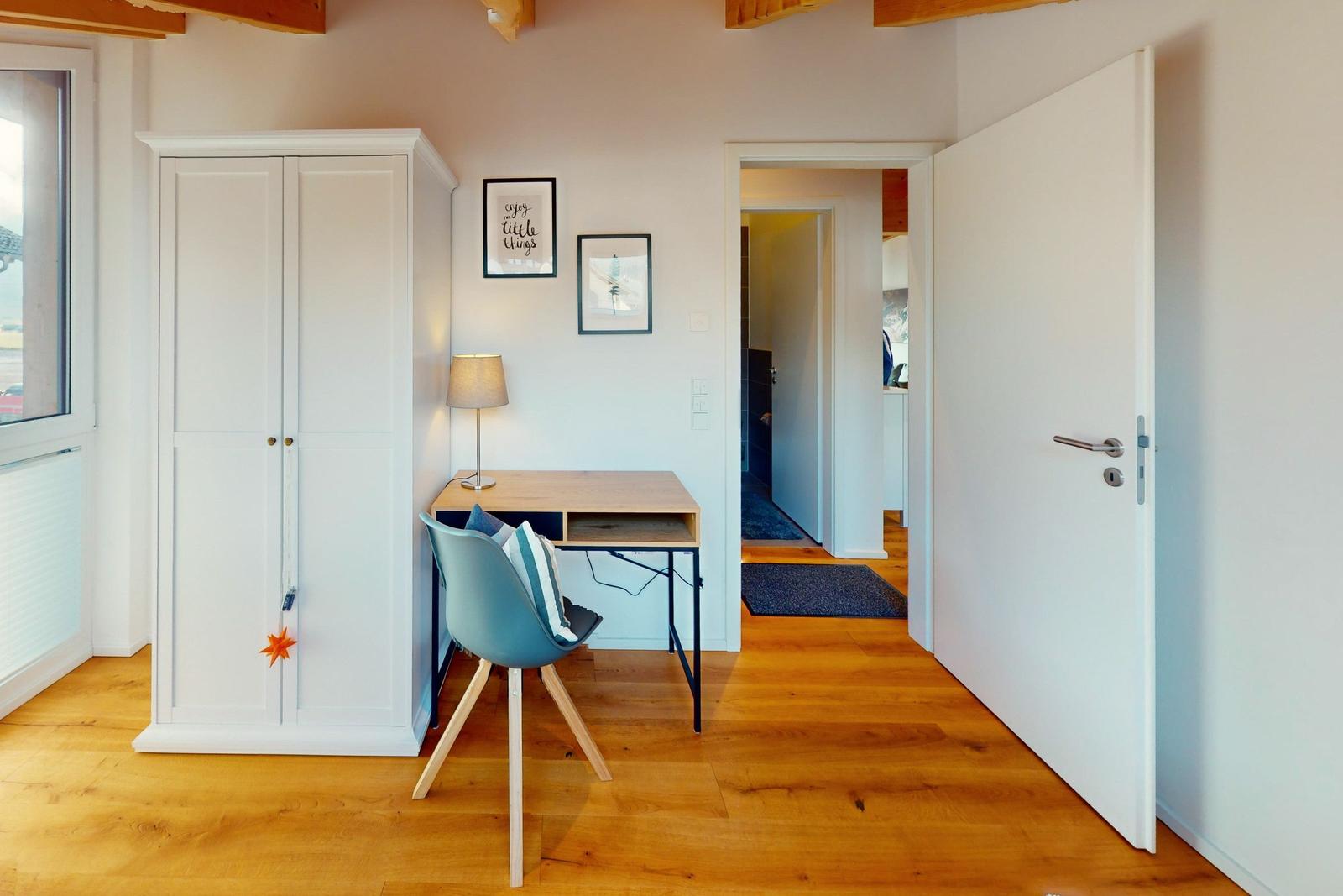Ein Raum mit Schreibtisch, Stuhl und Kleiderschrank. Holzboden und weiße Wände.