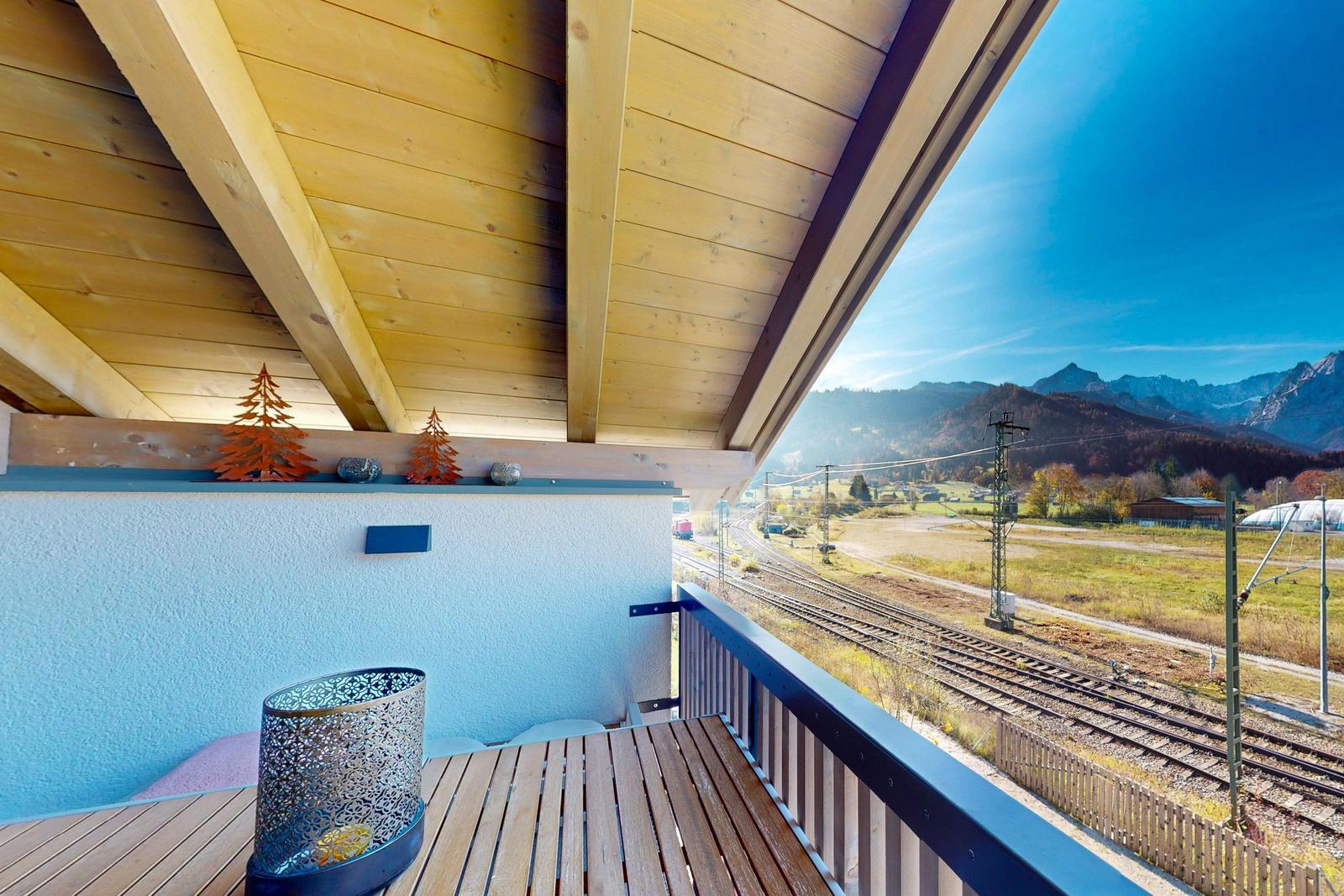 Terrasse mit Holzdecke und Tisch, Blick auf Bahngleise und Berge.