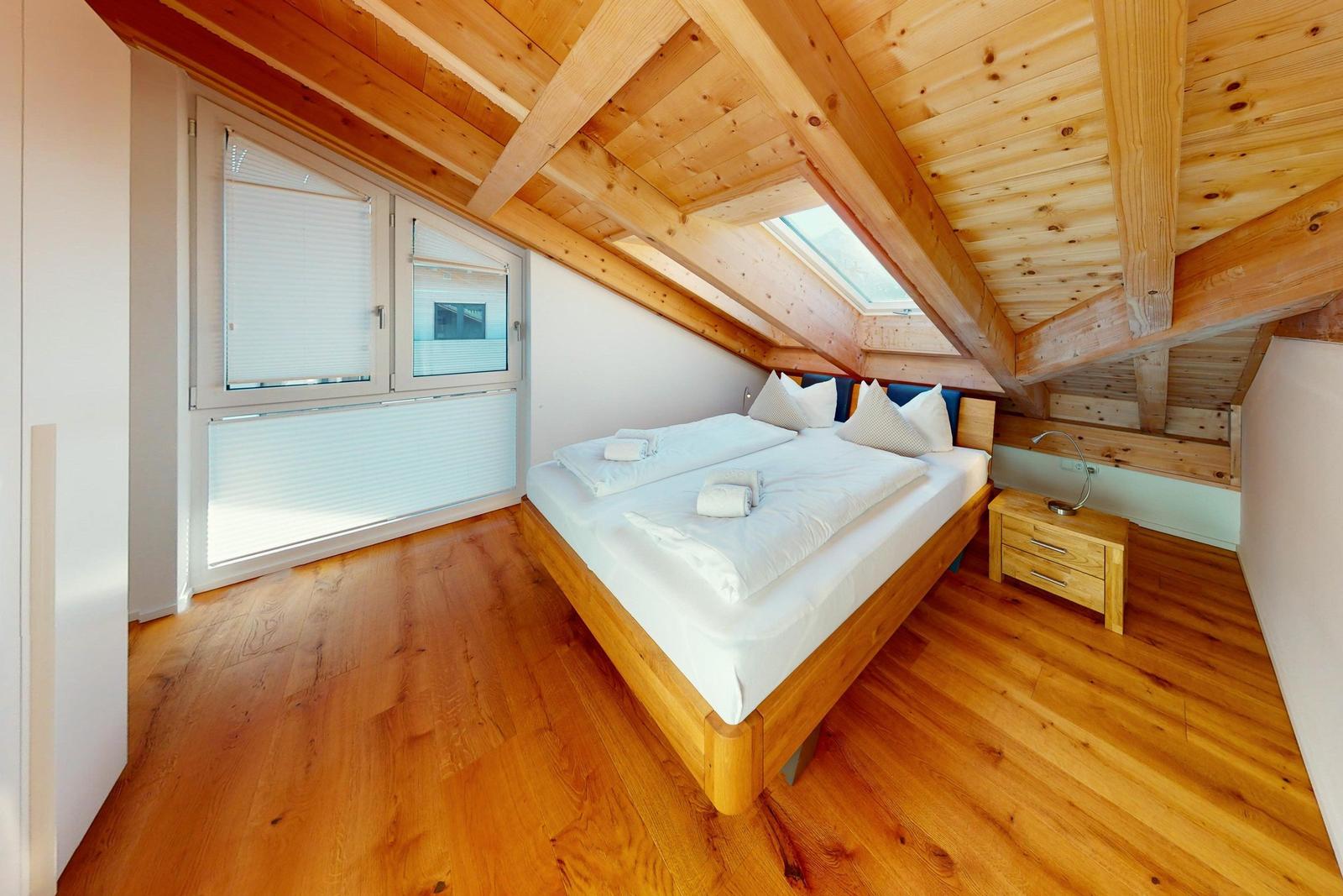 Schlafzimmer mit Holzboden, Dachschrägen und Fenster. Bett mit weißer Bettwäsche und Nachttisch.