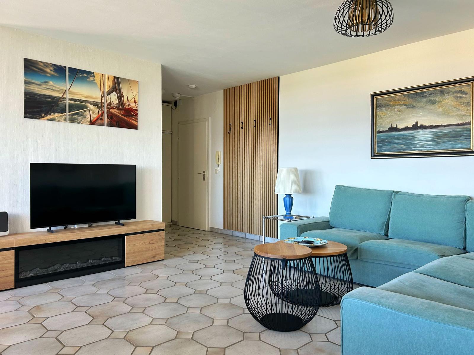 Wohnzimmer mit blauem Sofa, Fernseher und Holzregal. Bodenfliesen und Wandbilder.