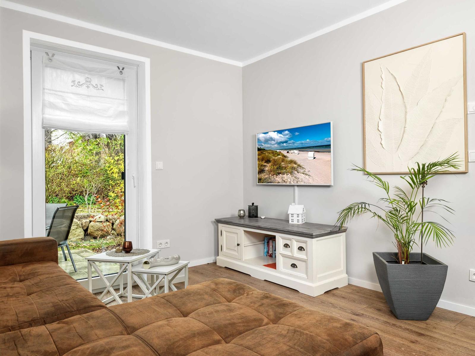 Wohnzimmer mit braunem Sofa, Fernseher und Pflanze. Fenster zeigt Garten.