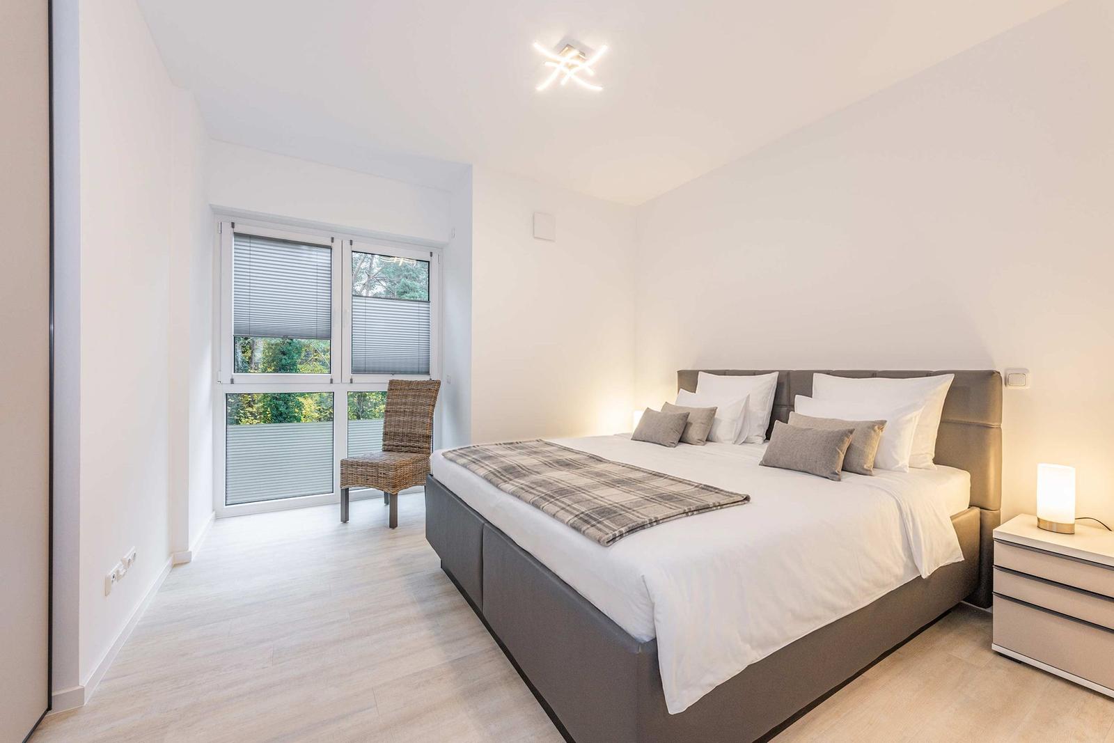 Schlafzimmer mit Doppelbett, Nachttisch und Stuhl vor Fenster mit Blick auf Grün.