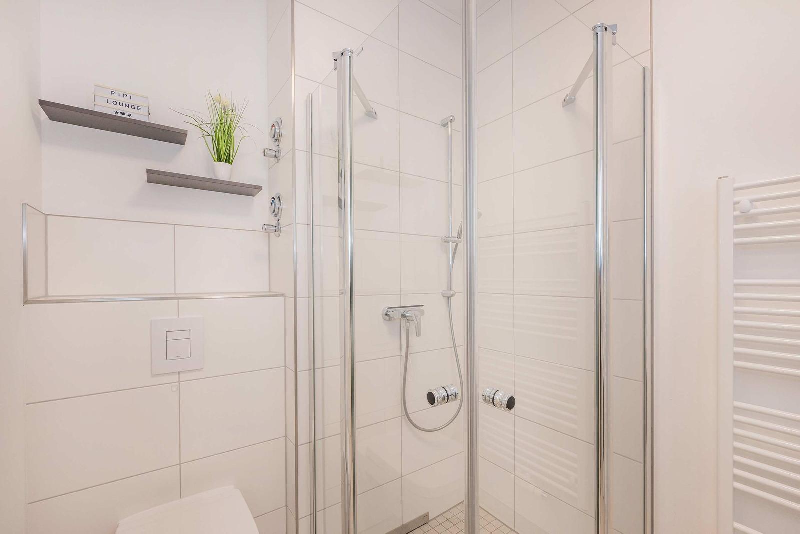 Dusche mit Glaswand, Duschbrause und Heizung. Wandregal mit Pflanze und Schild.
