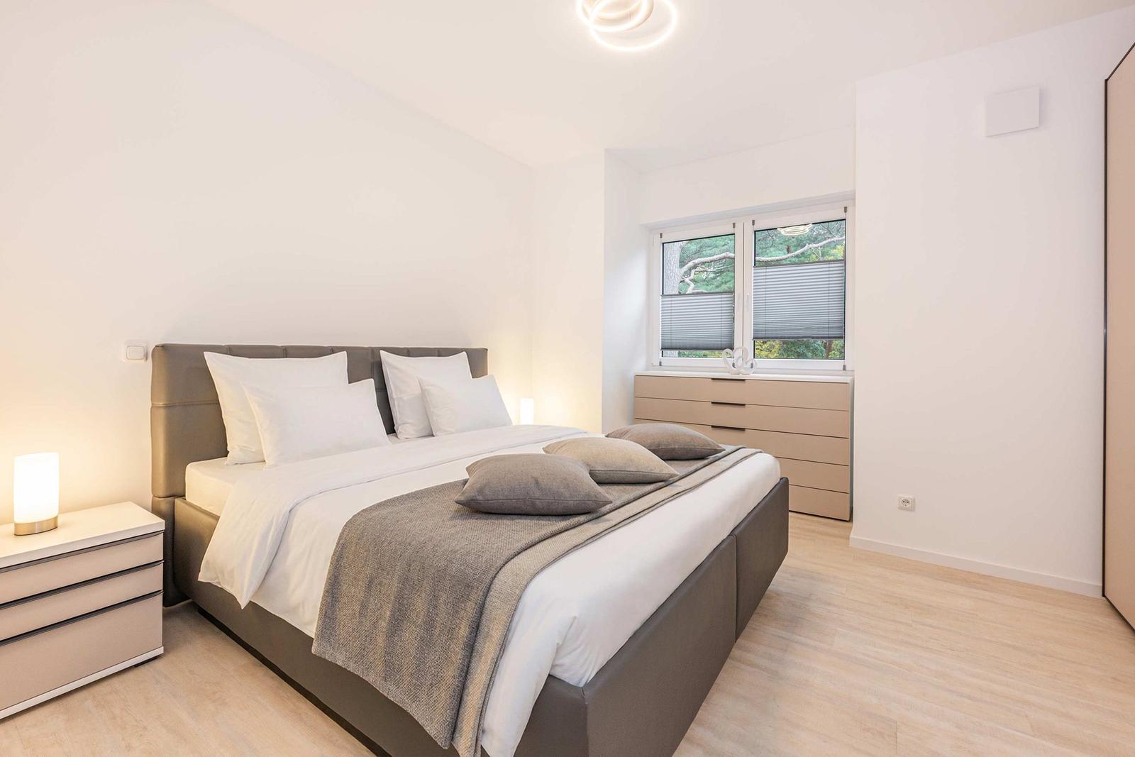 Schlafzimmer mit Bett, Nachttisch und Fenster. Holzboden und neutrale Farben.