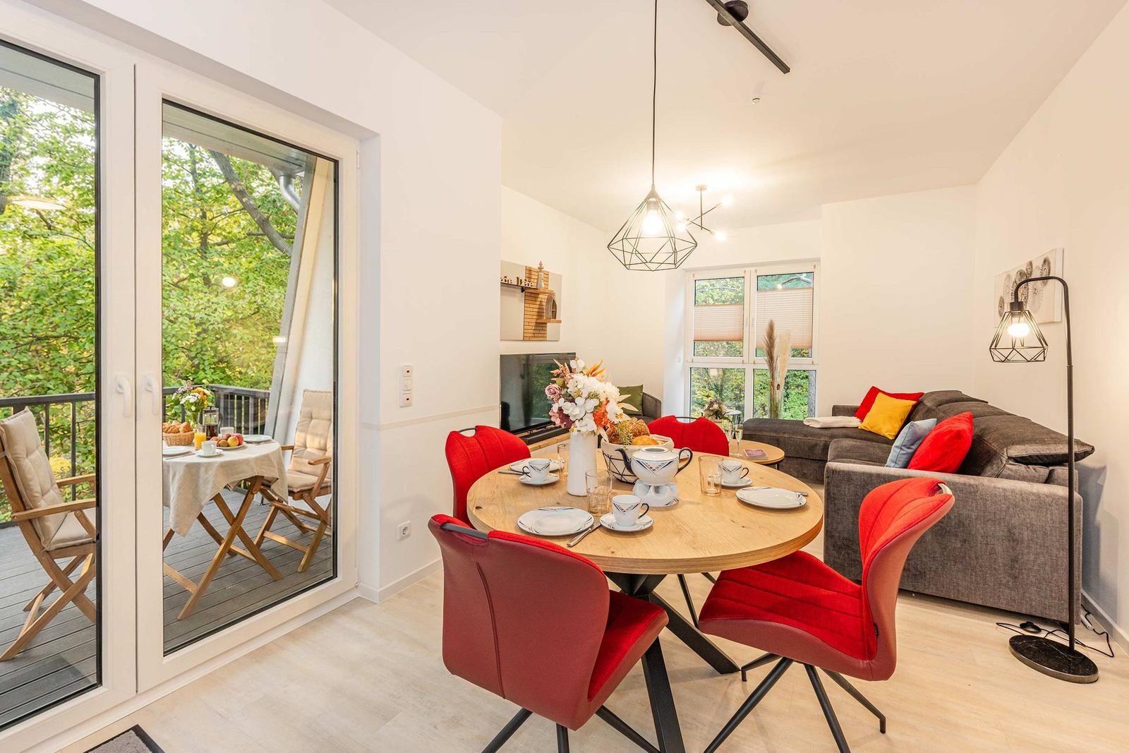 Wohn- und Essbereich mit runden Tisch, roten Stühlen und Balkon mit Blick auf Grün.
