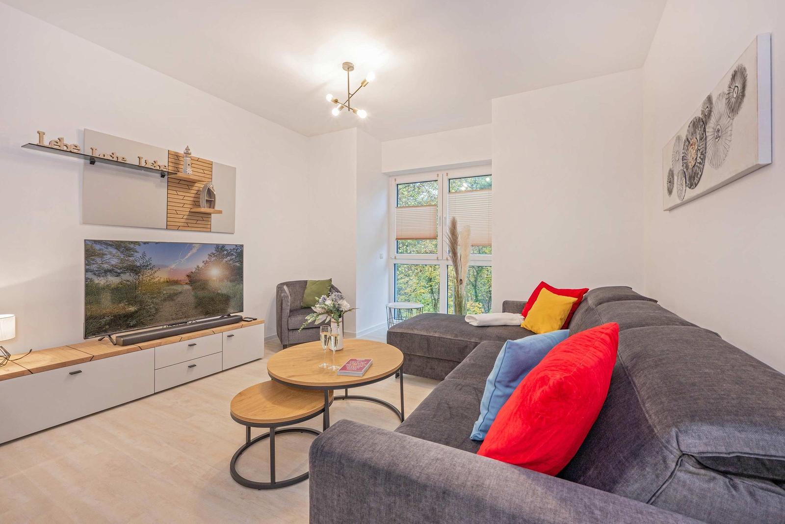 Wohnzimmer mit Sofa, Fernseher und Tisch. Fenster mit Blick auf Grün.