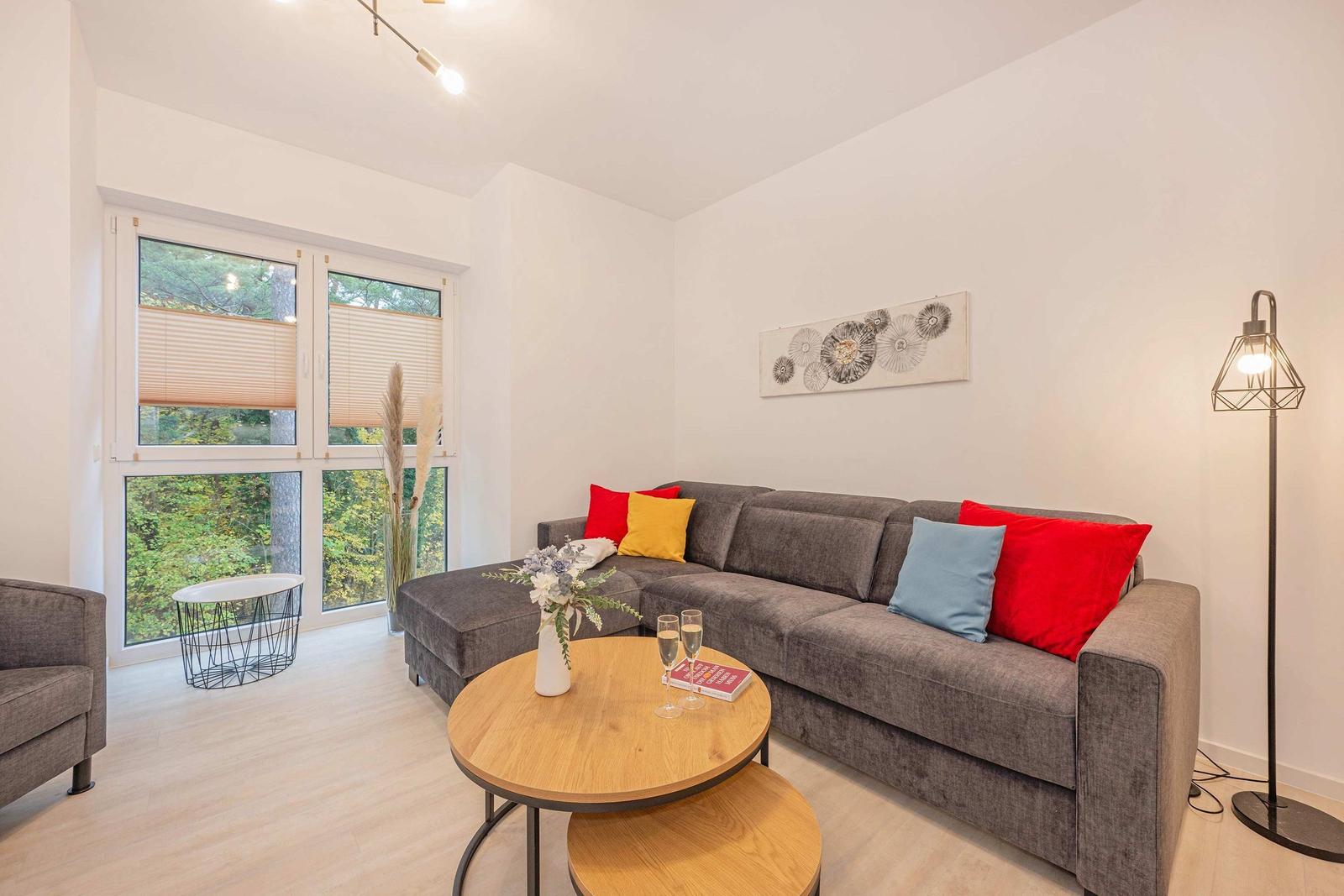 Wohnzimmer mit grauem Sofa, runden Tischen und Blick auf Wald.