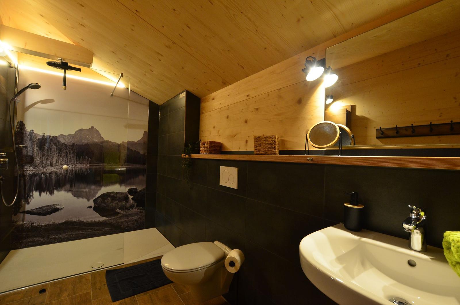Badezimmer mit Dusche, Waschbecken und Toilette. Holzwände und dunkle Fliesen.