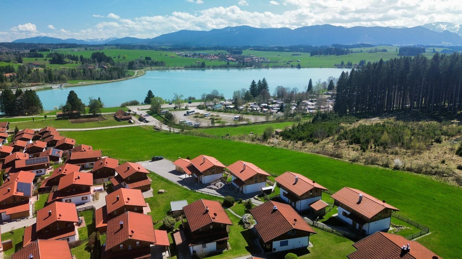 Häuser mit roten Dächern liegen an einem See, umgeben von Wiesen und Bergen.