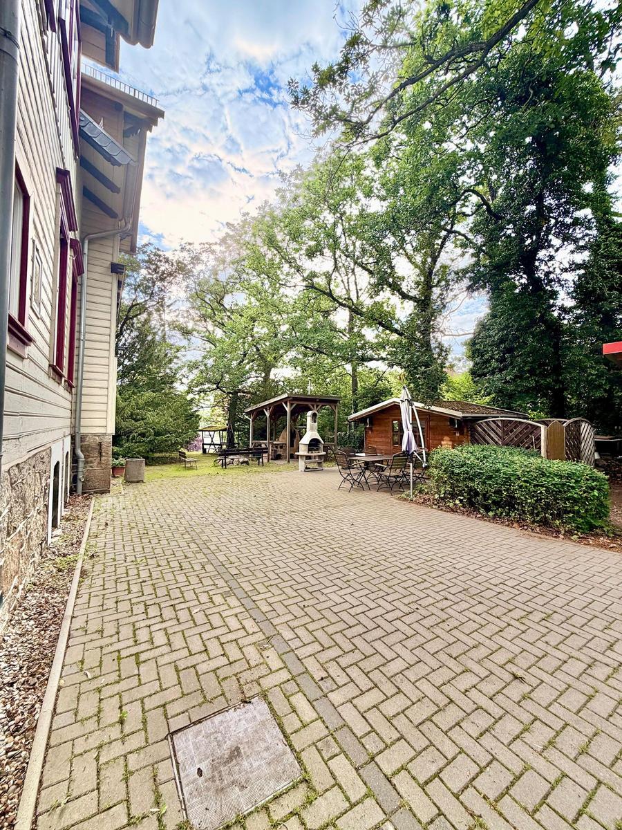 Gepflasterter Hof mit Grill, Tisch und Bäumen. Holzhaus und Pavillon im Hintergrund.
