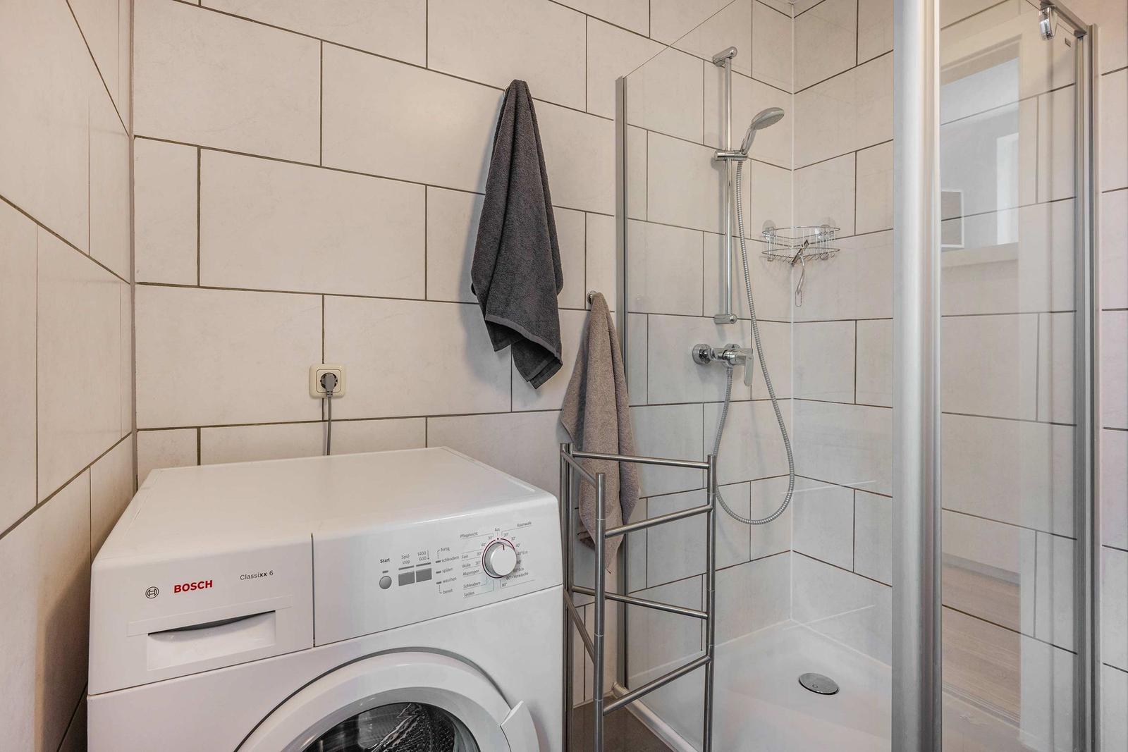 Waschmaschine und Dusche mit Glaswand in einem Badezimmer mit Fliesenwand.