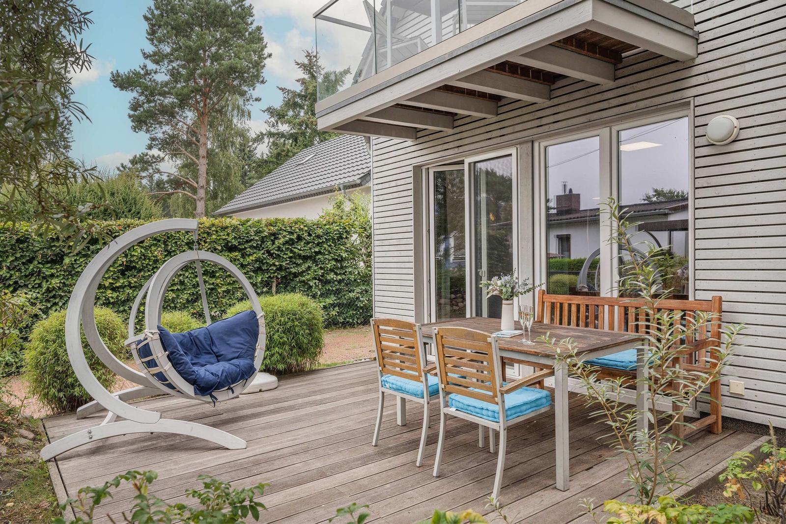 Terrasse mit Holztisch, Stühlen und Hängesessel. Moderne Holzhausfassade und grüner Garten.