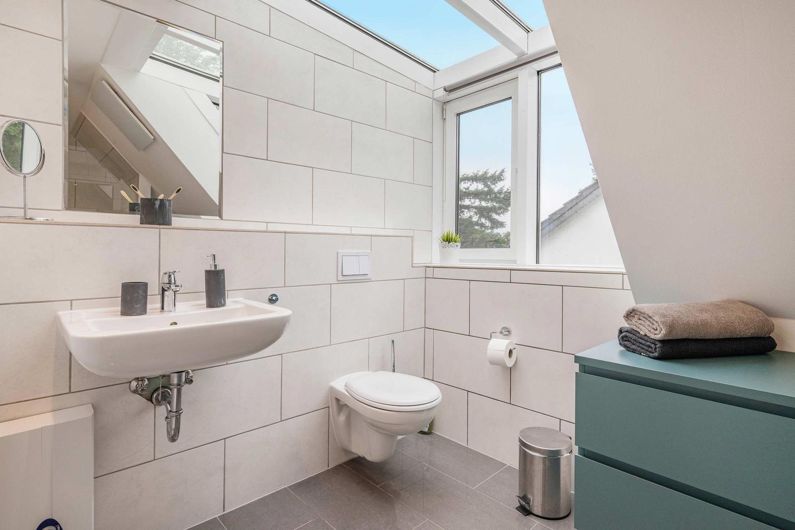 Badezimmer mit Waschbecken, Toilette und Fenster. Auf einem Schrank liegen Handtücher.
