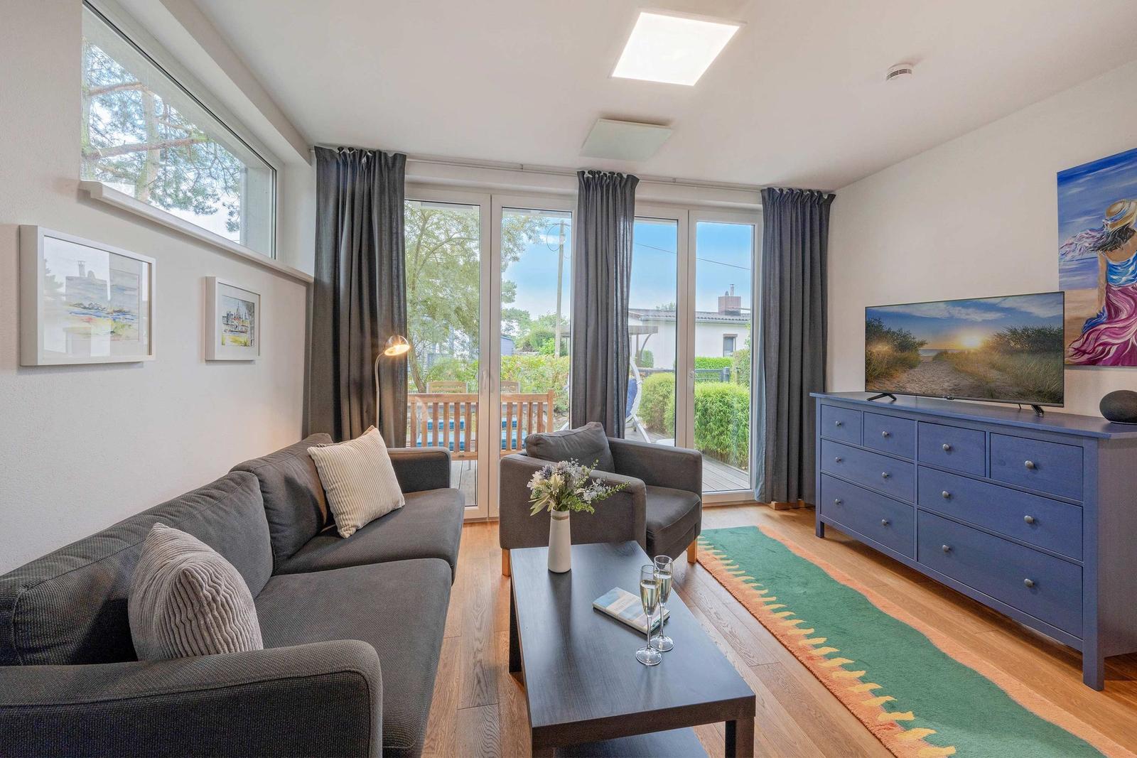 Wohnzimmer mit Sofa, Sessel, Couchtisch und TV auf blauem Schrank. Glastür führt auf Balkon.