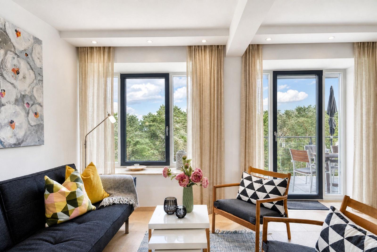 Wohnzimmer mit Sofa, Sessel und Balkonblick. Fenster und Türen mit Vorhängen.