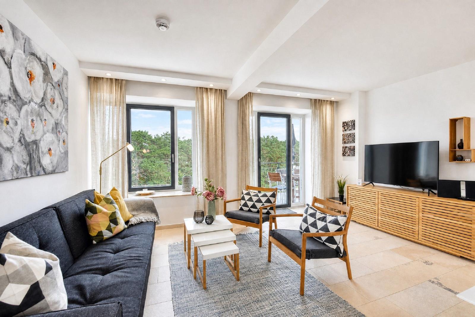 Wohnzimmer mit Sofa, zwei Stühlen, TV und Blick auf Balkon mit Blick auf Wald.