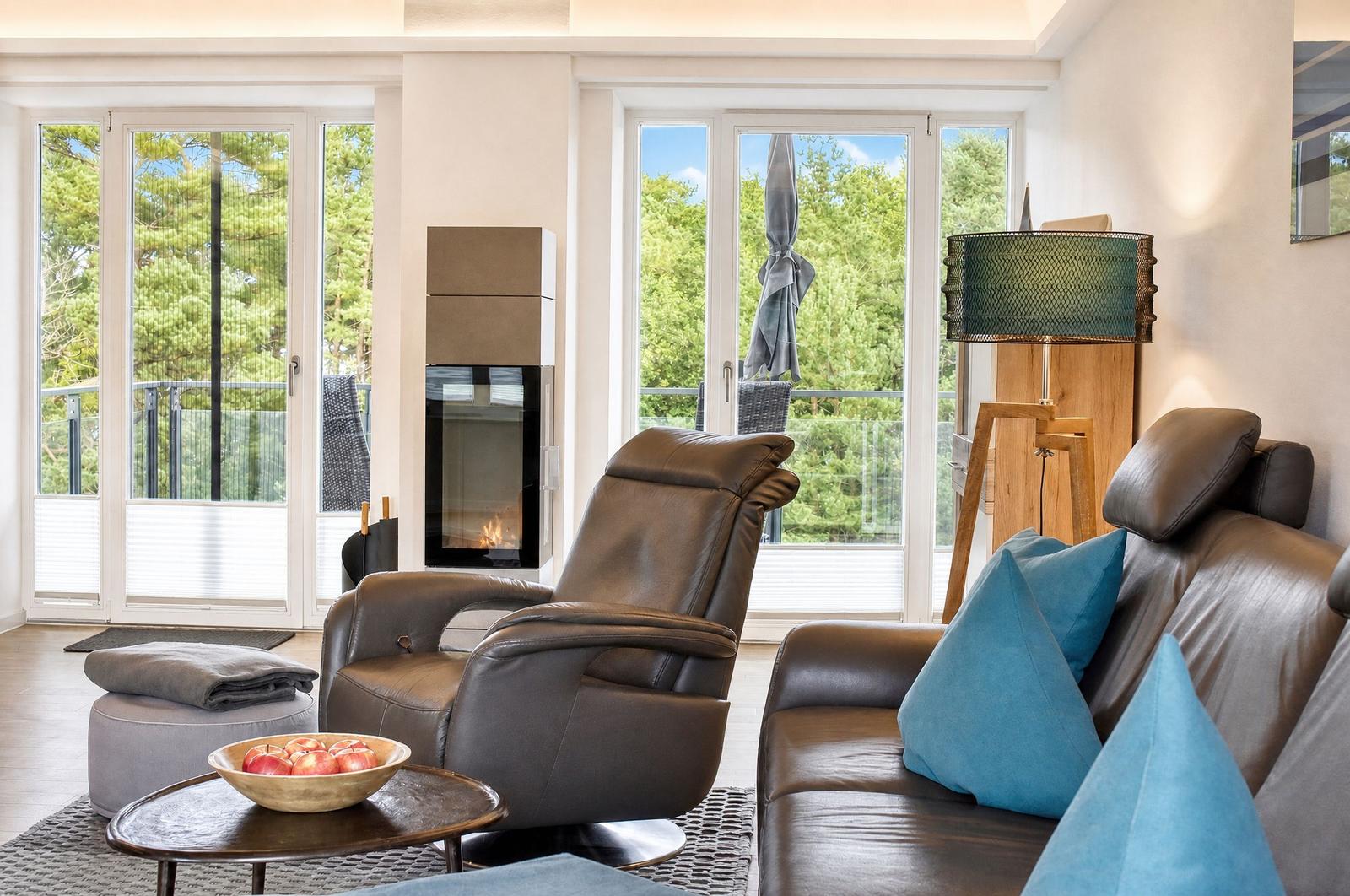 Wohnzimmer mit Ledersofa, Sessel und Kamin. Große Fenster mit Blick auf Garten.