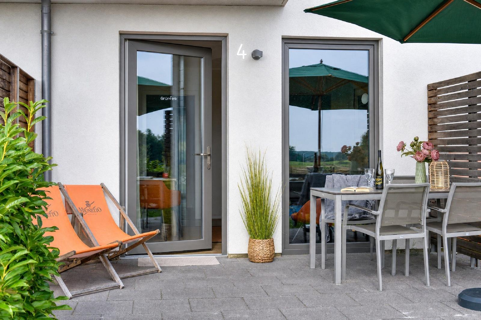 Terrasse mit Tisch, Stühlen, Sonnenschirm und Liegestühlen. Eingangstür und Pflanze sind sichtbar.