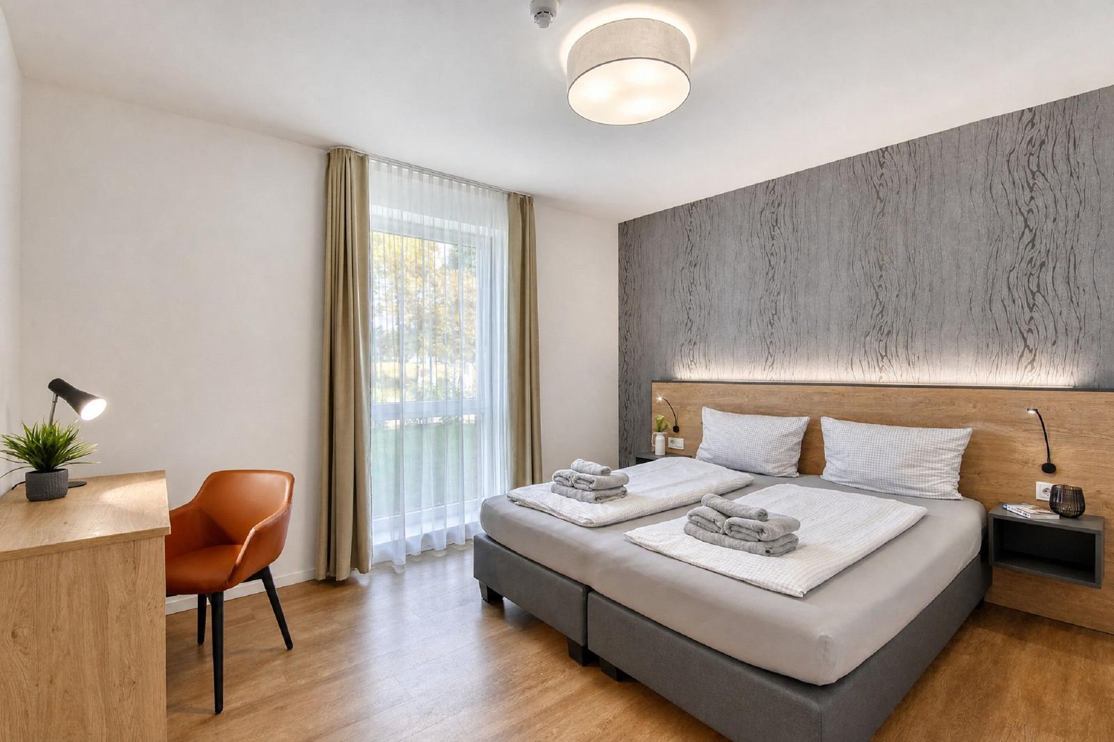 Schlafzimmer mit Doppelbett, Schreibtisch und Fenster. Wand mit Holzoptik.