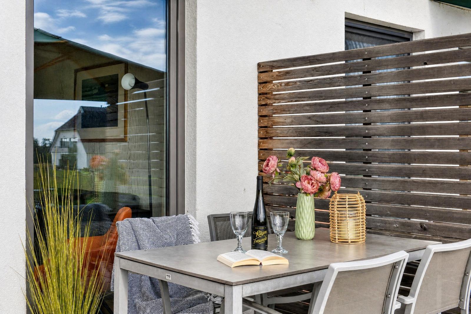 Terrasse mit Tisch, Stühlen, Blumen und Weinflasche. Holzgeländer und Fenster im Hintergrund.