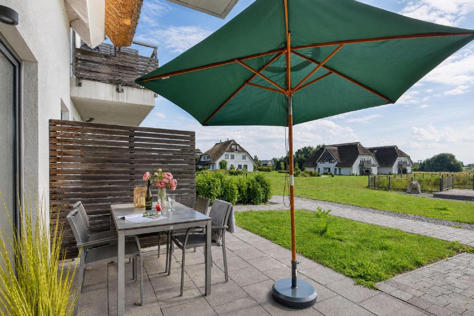 Terrasse mit Tisch, Stühlen und grünem Sonnenschirm. Blick auf grüne Wiesen und Häuser.