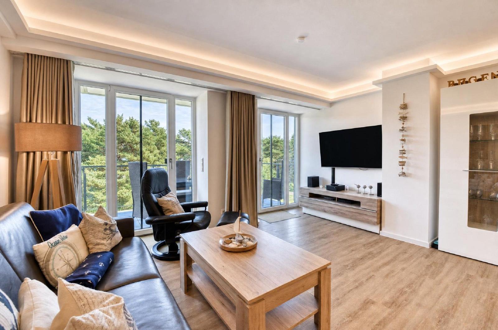 Wohnzimmer mit Sofa, Couchtisch und TV. Große Fenster mit Blick auf Balkon und Bäume.