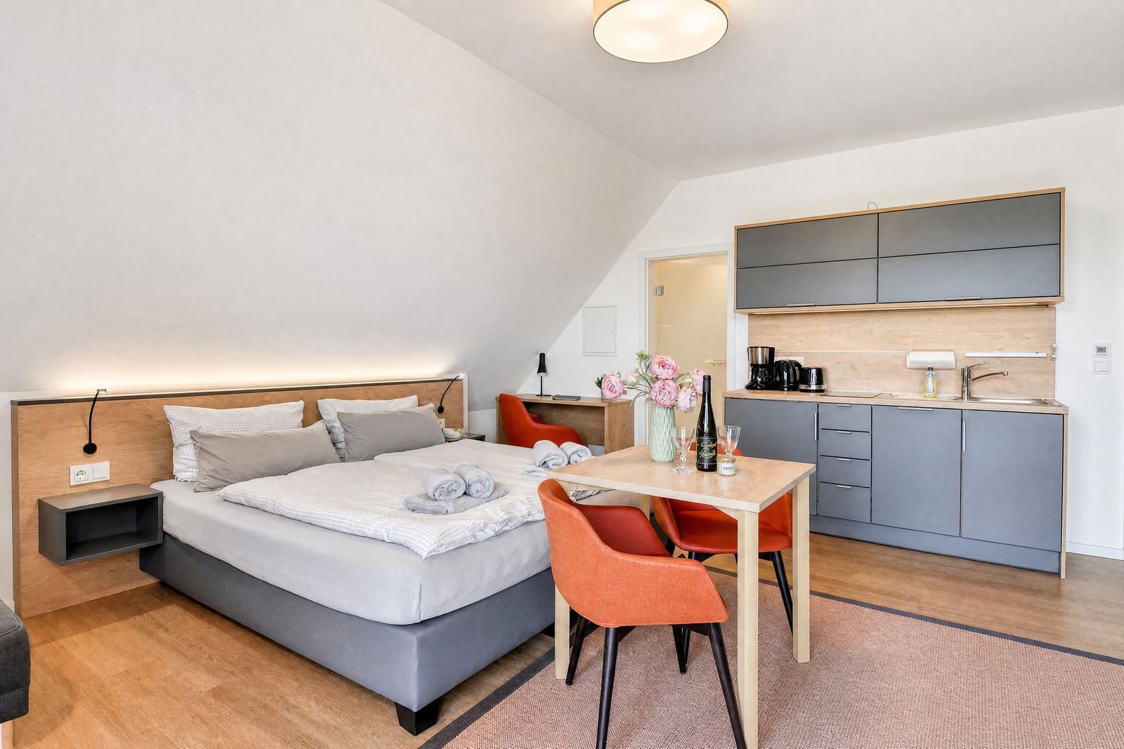 Ein Schlafzimmer mit Doppelbett, Küchenzeile und Esstisch mit zwei orangefarbenen Stühlen.