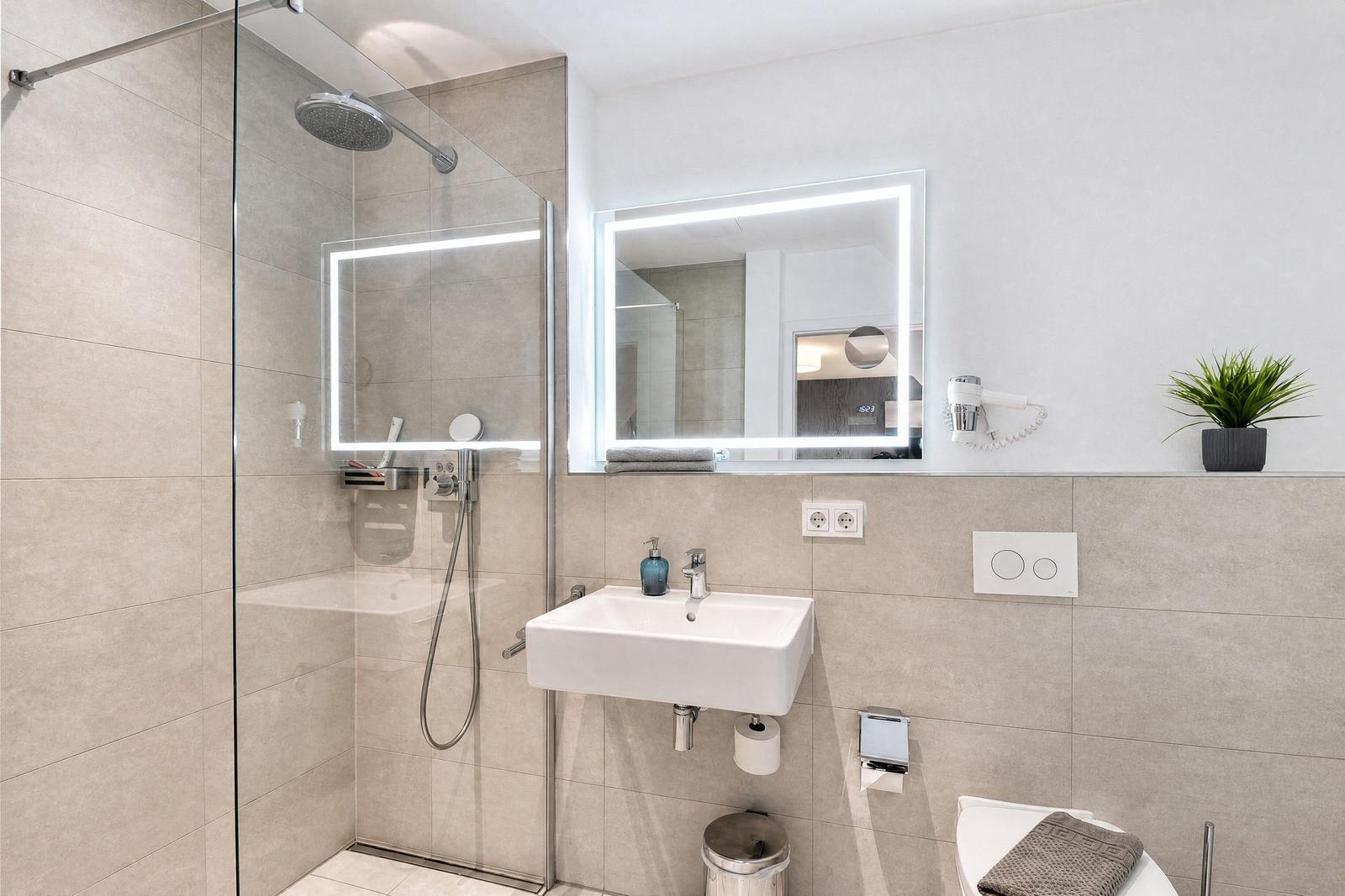 Bathroom with shower, sink, and toilet. Mirror with lighting and plant.