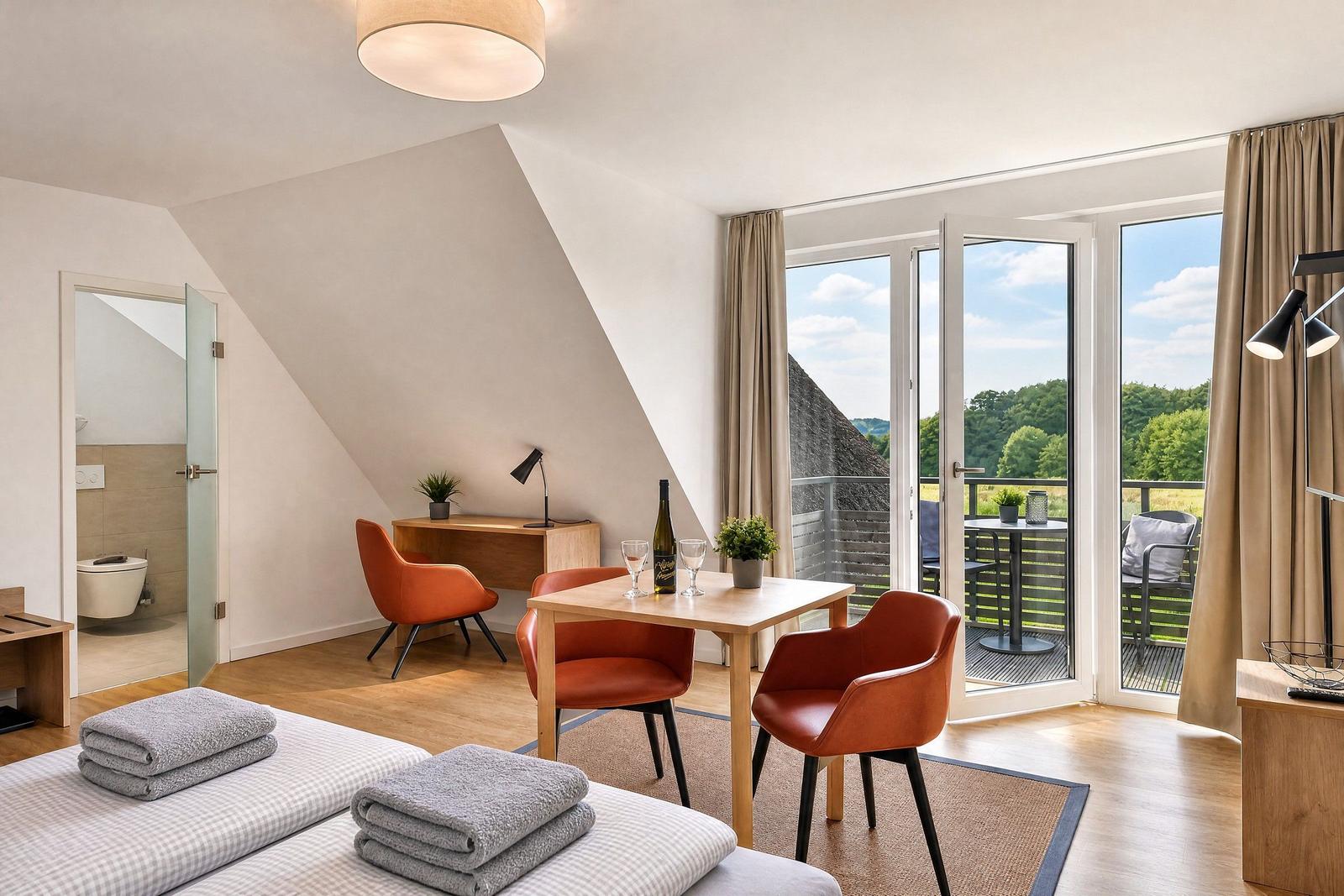 Room with balcony, table, chairs, and bathroom. View of green landscape.