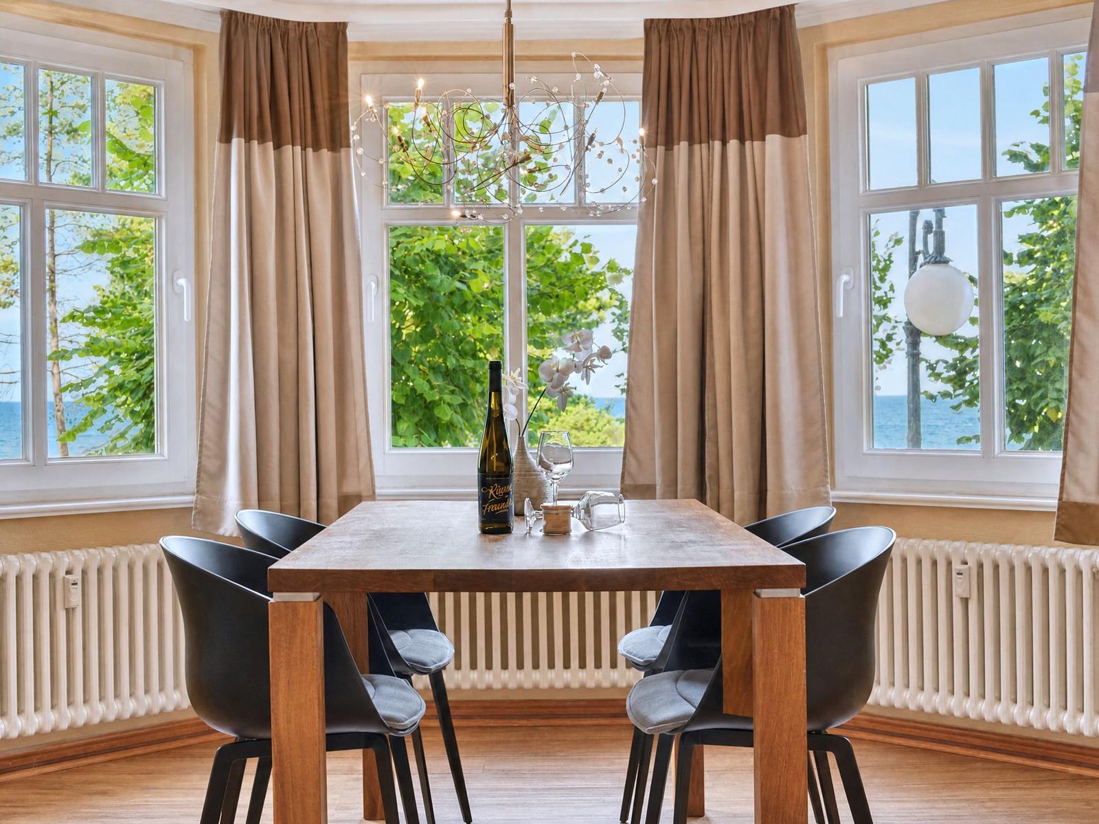 Esstisch mit sechs Stühlen vor großen Fenstern mit Blick auf Wasser und Bäume.