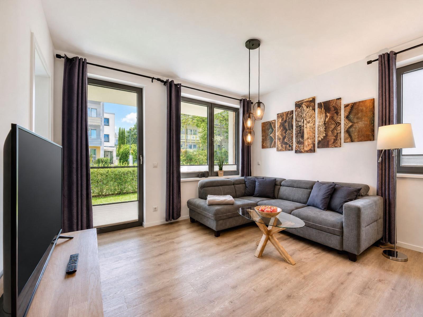 Living room with sofa, glass table, and TV. Large windows with garden view.