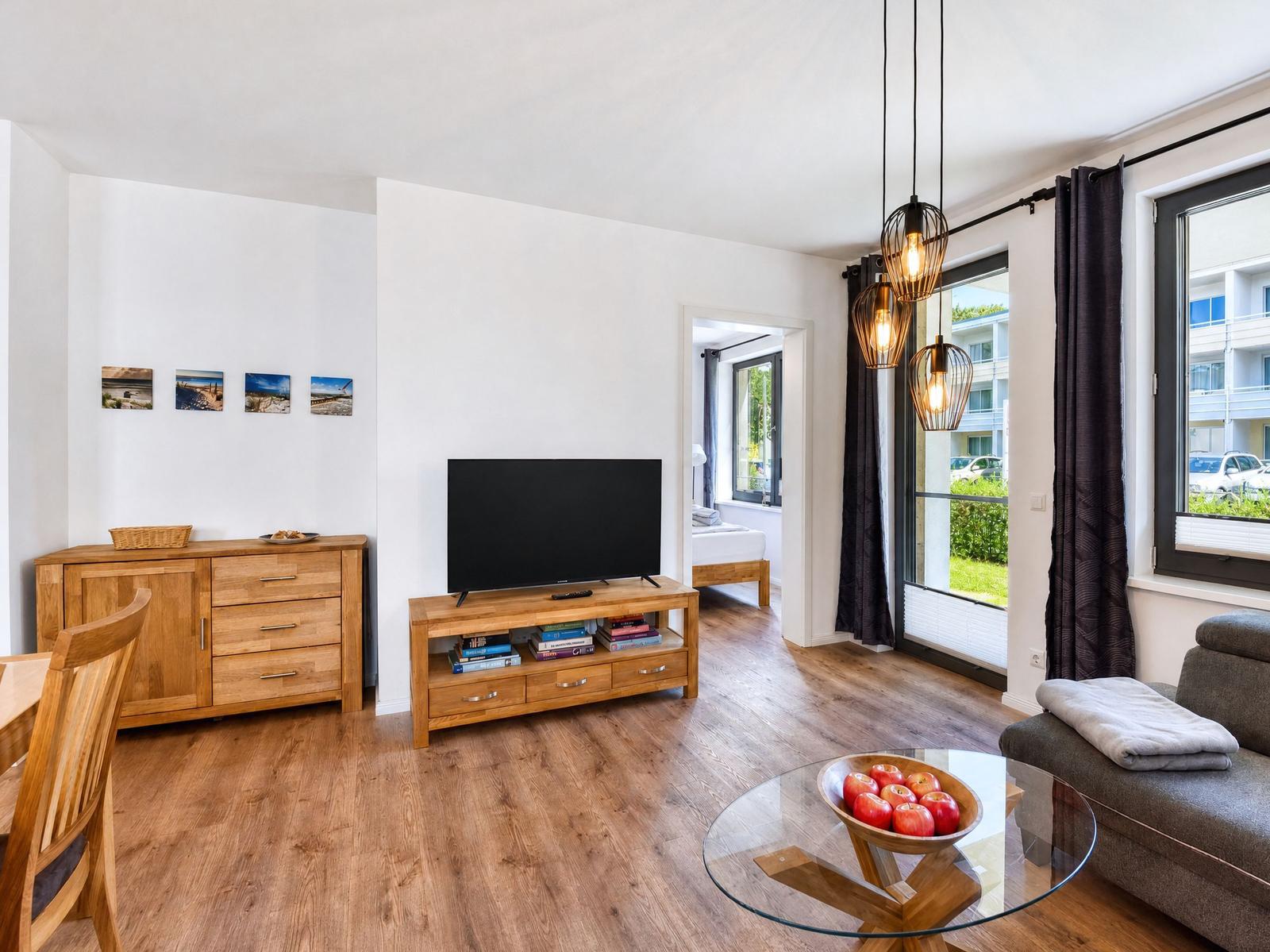 Living room with TV, glass table, and seating. Wooden floor and windows with view.