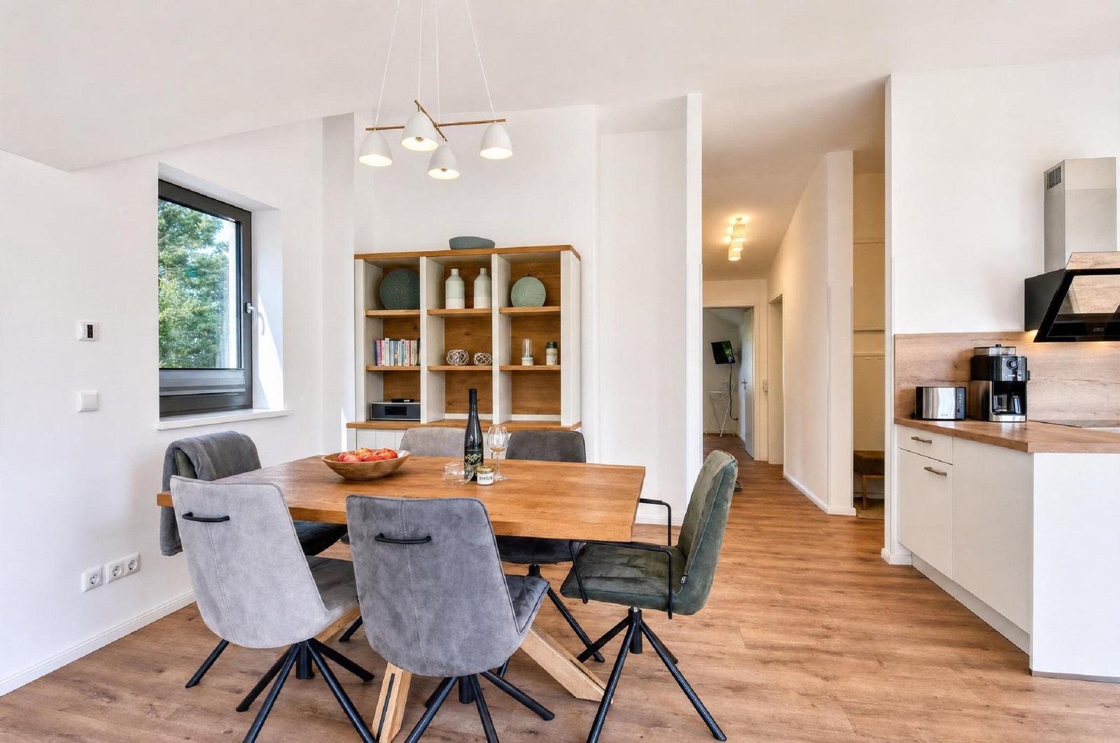 Essbereich mit Holztisch, Stühlen und Küchenbereich. Fenster mit Blick auf Grün.