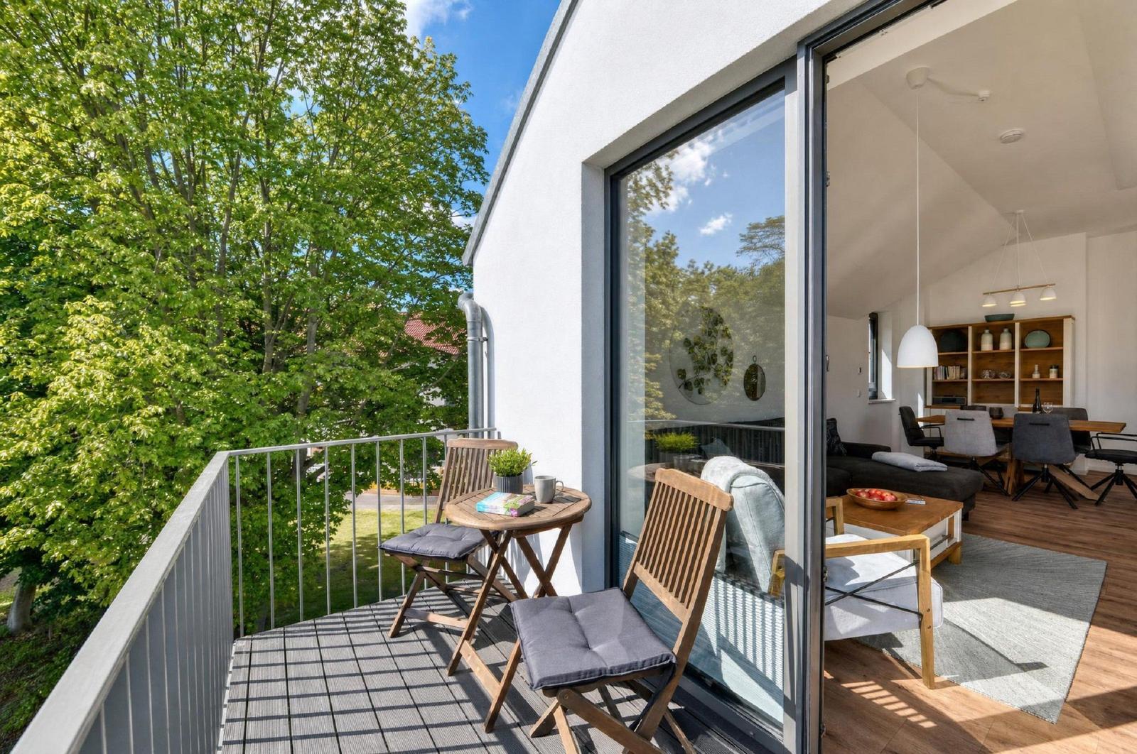 Terrasse mit Tisch und Stühlen, Blick auf grüne Bäume. Durchgehendes Glas führt in Wohnbereich mit Sofa und Tisch.