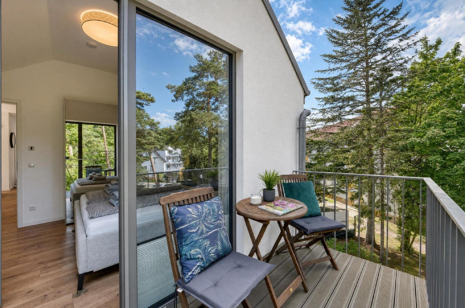 Terrasse mit zwei Stühlen und Tisch. Blick auf Bäume und Himmel. Im Hintergrund das Innere der Wohnung.