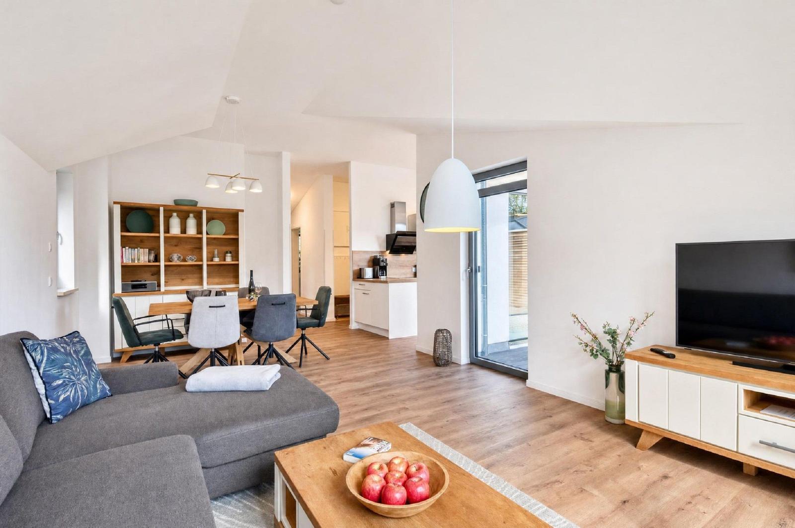 Wohnzimmer mit Sofa, Couchtisch und Fernseher. Küche und Essbereich im Hintergrund.