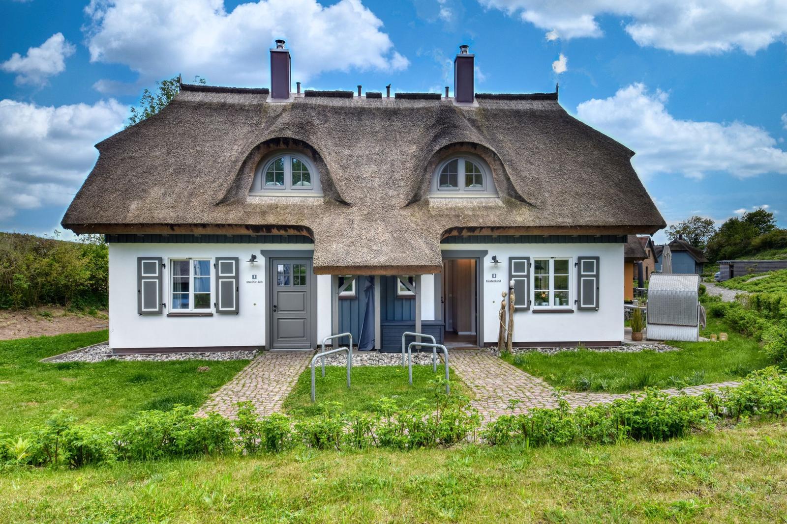 A house with thatched roof, white walls, and gray shutters. A paved path leads to the entrance.