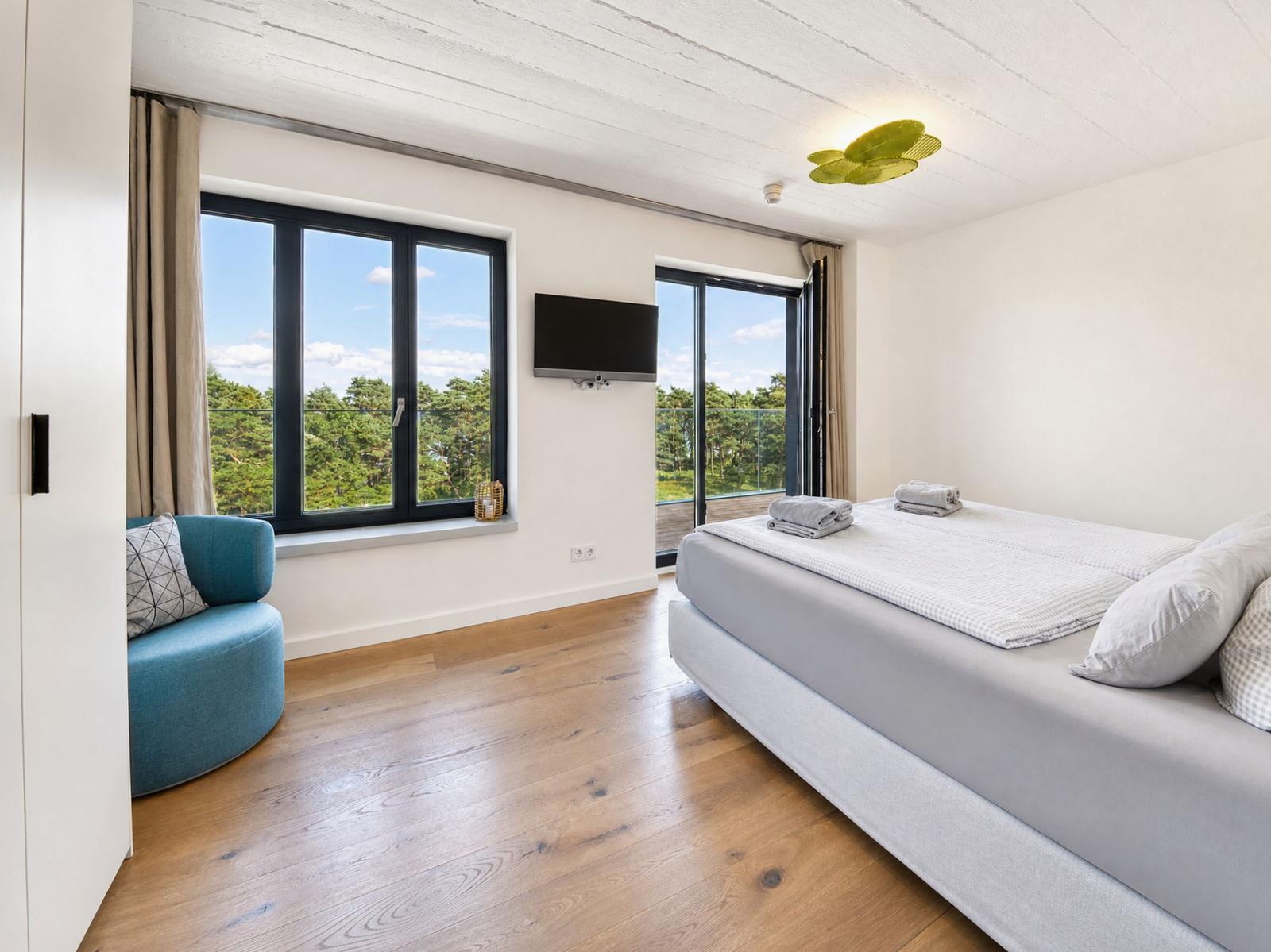 Schlafzimmer mit großem Bett, Fenster und Balkon mit Blick auf Wald.