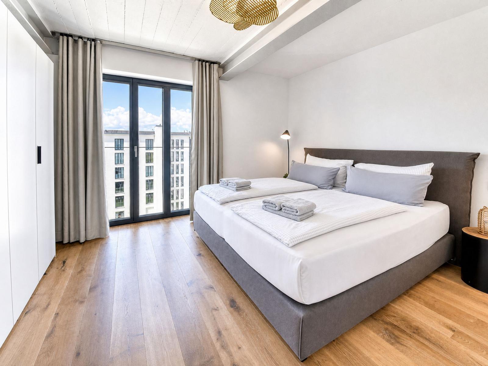 Schlafzimmer mit Doppelbett, Holzboden und Balkontür. Helle Wände und goldener Deckenleuchte.