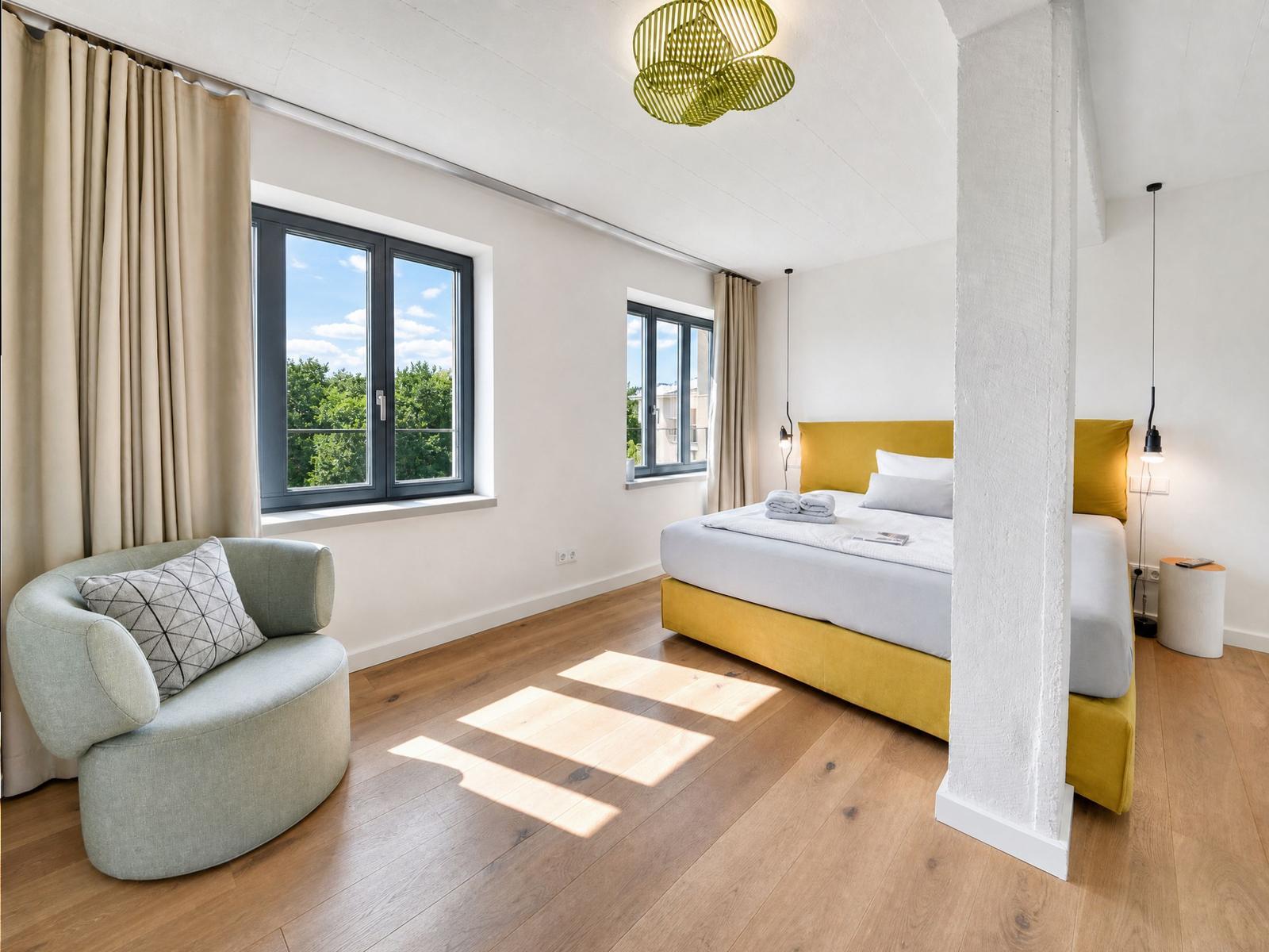 Schlafzimmer mit gelbem Bett, Holzboden und großem Fenster mit Blick auf Bäume.