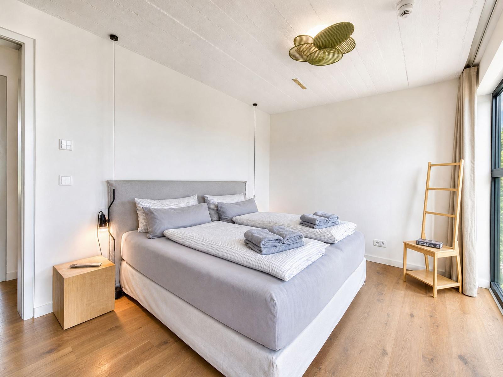 Ein Schlafzimmer mit Bett, Nachttisch und Kleiderständer. Holzboden und großes Fenster.